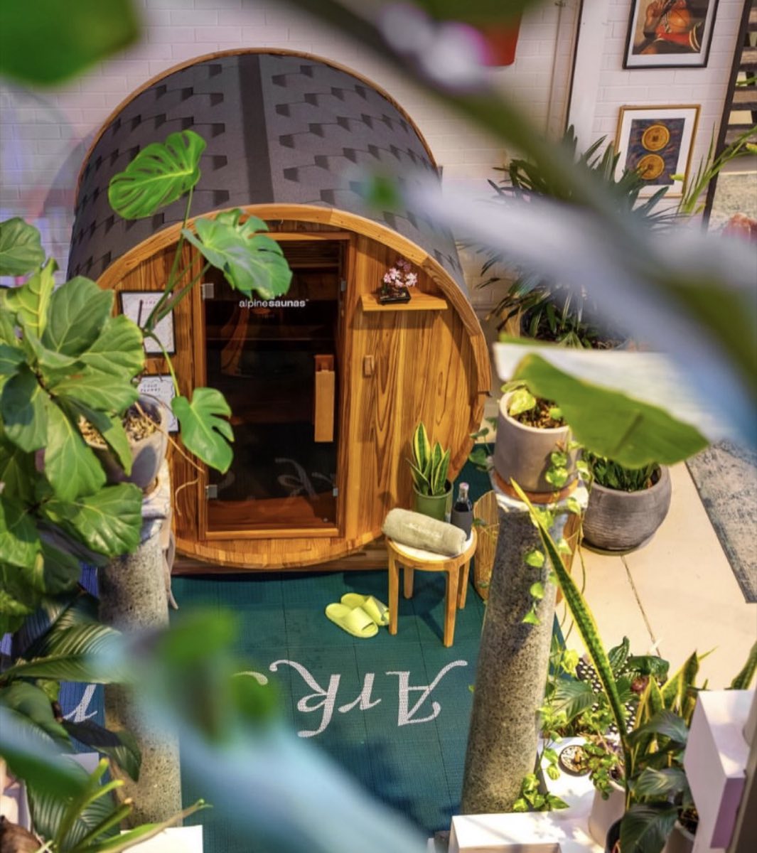 A barrel sauna framed by leaves, with the Ark Sports Club script on the wall behind